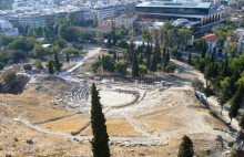 А здесь мы видим Театр Диониса. Культ Диониса появился в Афинах в шестом веке до н.э. Обряды, проходившие в честь этого бога, с необузданными песнями и плясками, тесно связаны с возникновением греческого театра. Начиная с шестого века до н.э. ритуал обогатился мимическими движениями и диалогами, вплоть до выхода на сцену актеров в театральных масках. В пятом веке до н.э. естественный уклон Акрополя решили использовать для сооружения театра. Сначала он был из дерева, но потом был снабжен каменными сидениями и постоянной сценой. Театр Диониса – это первый архитектурный пример той структуры театра, который стали применять в дальнейшем повсеместно. 