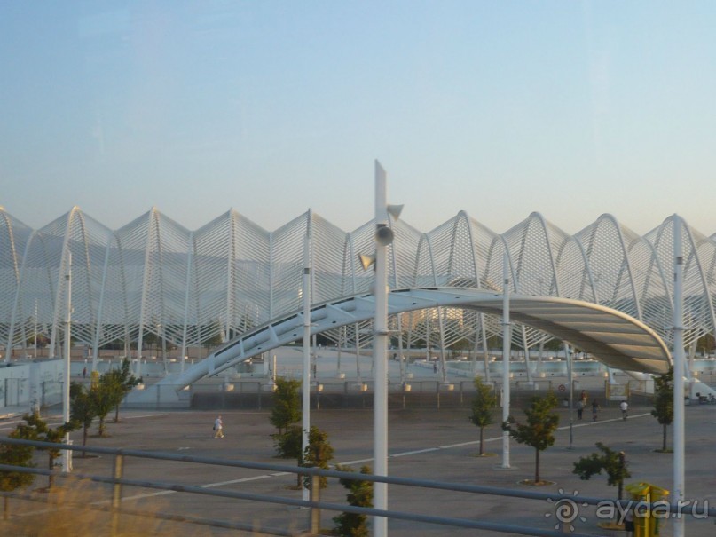 Олимпийский объект. Народ заходил под этот козырёк и дальше через коридор из железных «пальмовых ветвей» проходил на стадионы, треки и т.п. сооружения. Если внимательно приглядеться, на верхнем снимке видно, что по сторонам от коридора имеются бассейны, которые были возведены для охлаждения воздуха во время Олимпиады. Рассказывают, что в то время в Афинах стояла жуткая жара, и было немало желающих нырнуть в эти бассейны. Да только вот беда, глубина этих бассейнов была по колено. Пришлось в экстренном порядке расставлять кругом волонтеров, чтобы те не допускали таких заныров, а то и шею сломать недолго.    